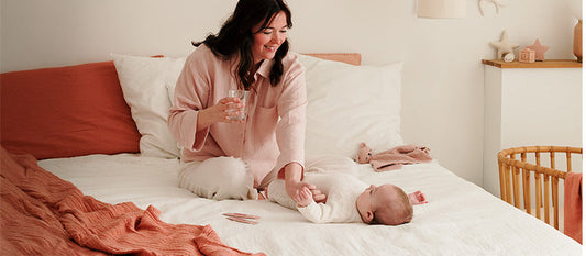 Une maman assise sur un lit joue avec son bébé qui est sur le dos.